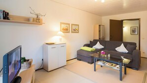 A modern living room with a flat-screen TV, a sofa, a coffee table, a small cabinet, and a lamp. - Apartment 'Bergfrieden' with Private Terrace, Shared Garden and Wi-Fi (Bad Herrenalb)
