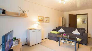 Ein modernes Wohnzimmer mit Flachbildfernseher, einer Couch, einem Couchtisch, einer kleinen Kommode und einer Lampe.