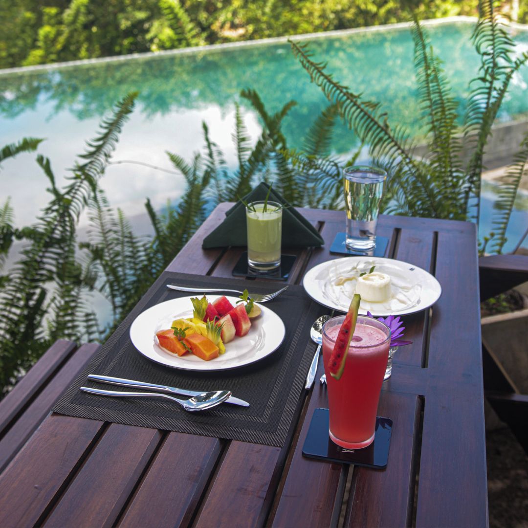 Una mesa de comedor con un plato de fruta, un vaso de bebida roja y un vaso de bebida verde, frente a un fondo de piscina y vegetación.
