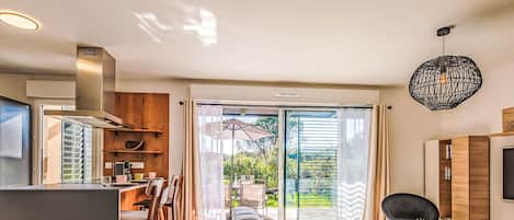A modern living room with a sofa, a dining area with chairs, a kitchen with a stove, and a large sliding glass door leading to a garden.