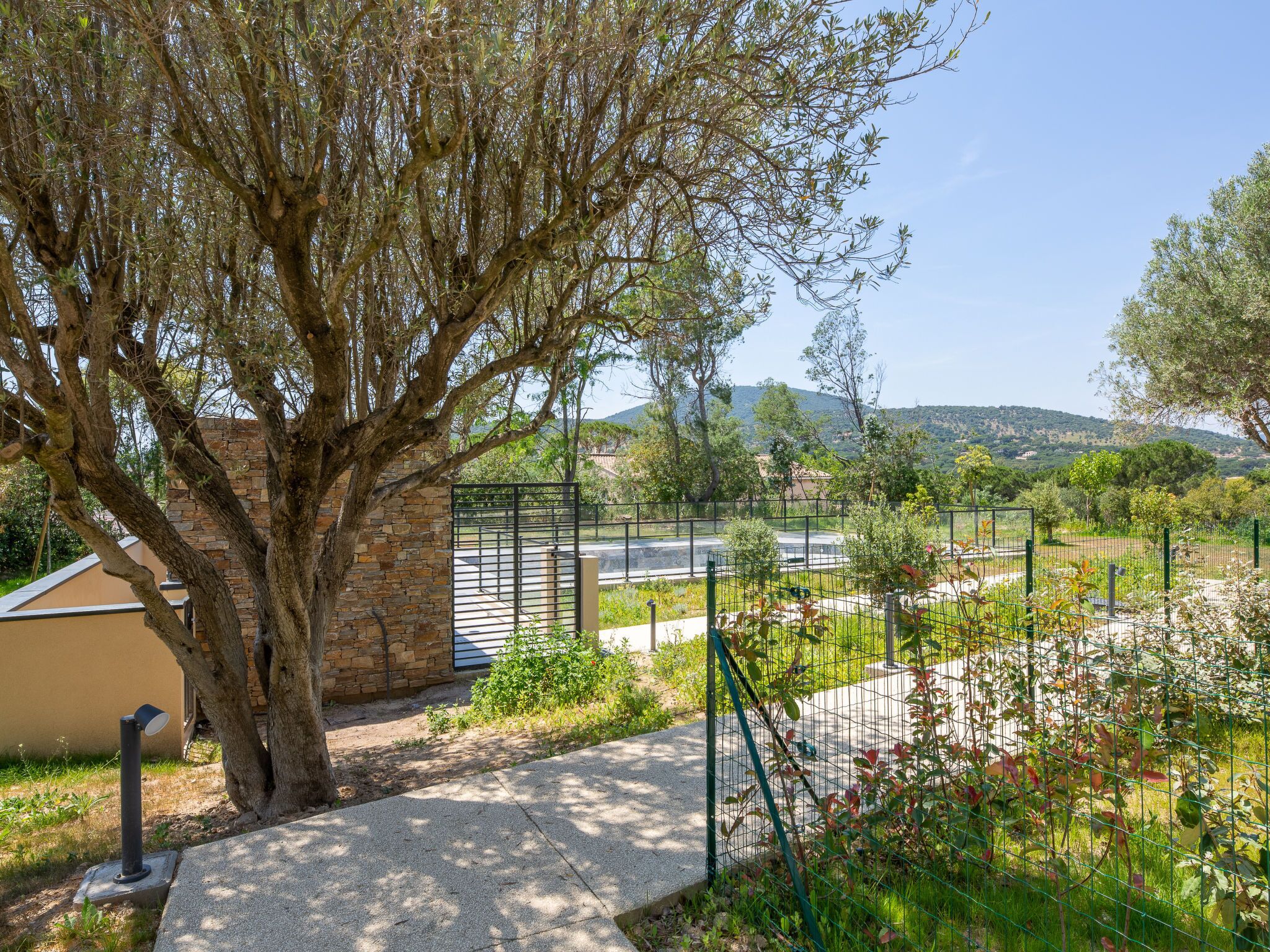 Un sendero bordeado de árboles que conduce a una entrada porticada, con un muro de piedra y una reja metálica.