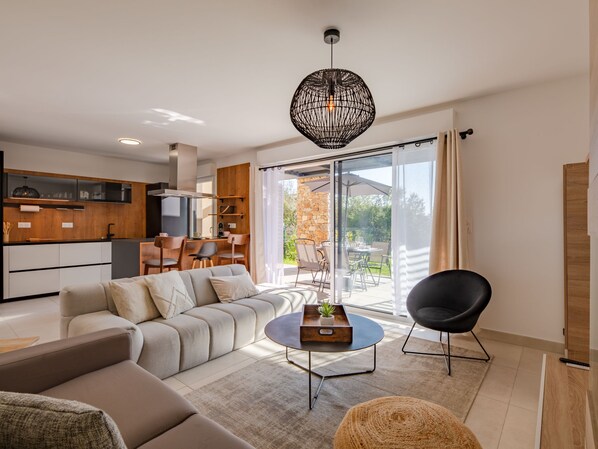 Un salon moderne avec un canapé, une table basse ronde et un fauteuil noir. On y trouve également un coin cuisine avec des meubles en bois et une table à manger avec des chaises. La pièce est dotée de grandes fenêtres et offre une vue sur l’extérieur.