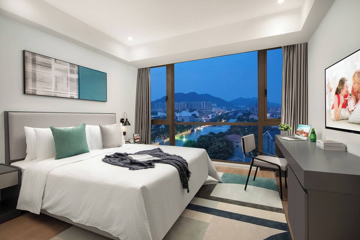 Una habitación de hotel moderna con una cama grande, un escritorio y vista a un paisaje urbano al anochecer.