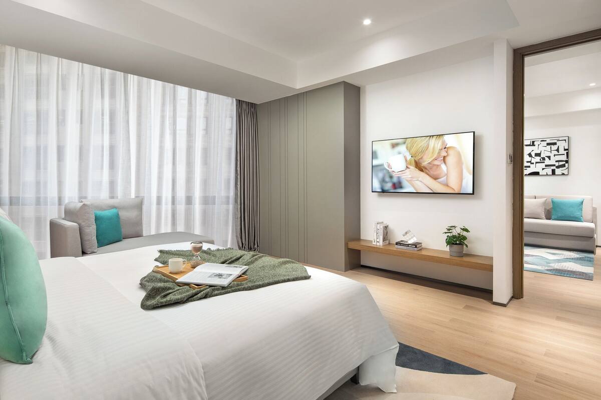 Una habitación de hotel moderna con una cama grande, un televisor montado en la pared, una mesita de noche con una planta y vistas a la ciudad a través de cortinas traslúcidas.