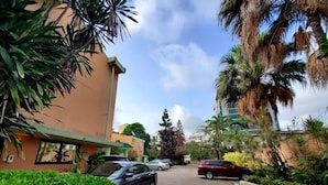 A street view with parked cars, a building with a green awning, and lush greenery including palm trees. - COLONADES HOTEL (Lagos)