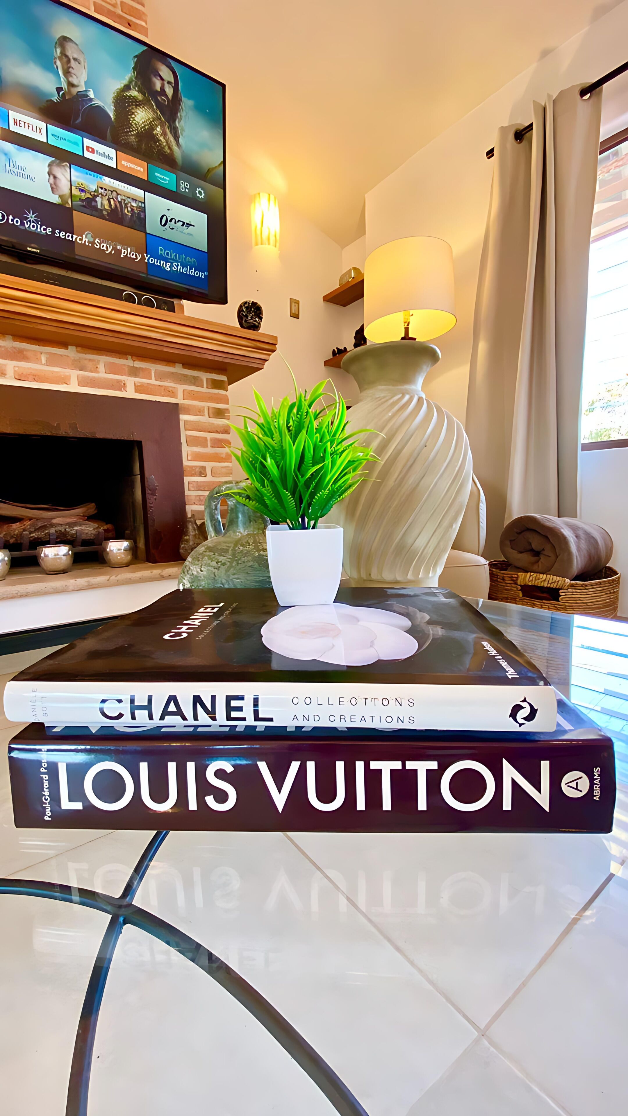 A living room with a TV displaying a movie, a fireplace, a lamp, a potted plant, and a stack of books including 'Chanel' and 'Louis Vuitton'.