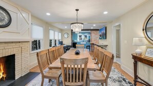 A dining area with a wooden table and chairs, a fireplace, and a kitchen in the background.