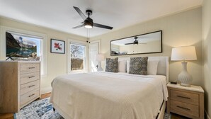 A bedroom with a large bed, a ceiling fan, a nightstand with a lamp, a dresser, and a window with a view of a mountain landscape.
