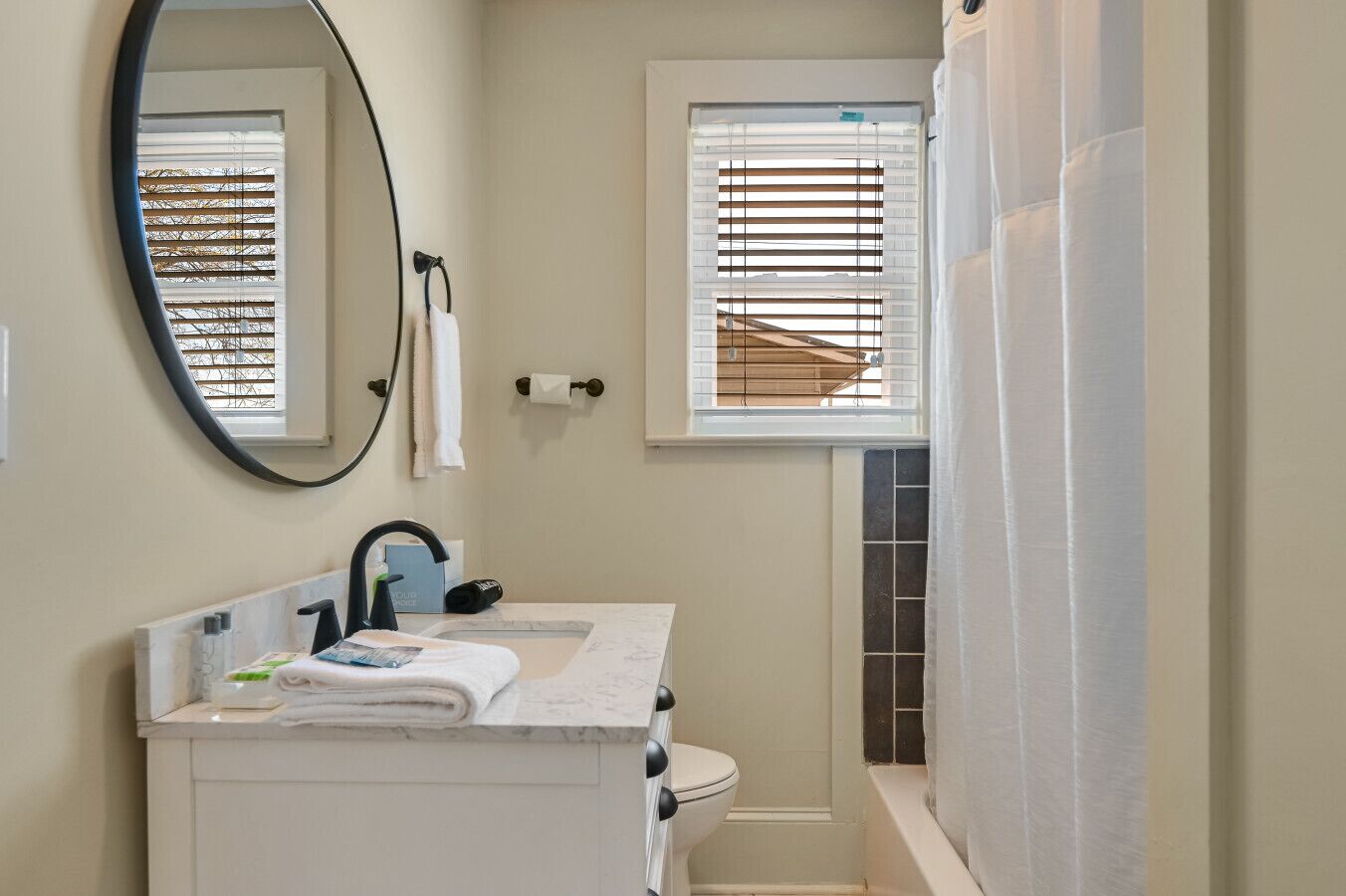 Une salle de bain équipée de toilettes, d’un lavabo, d’un miroir et d’une fenêtre avec des stores.