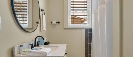 Une salle de bain équipée de toilettes, d’un lavabo, d’un miroir et d’une fenêtre avec des stores.