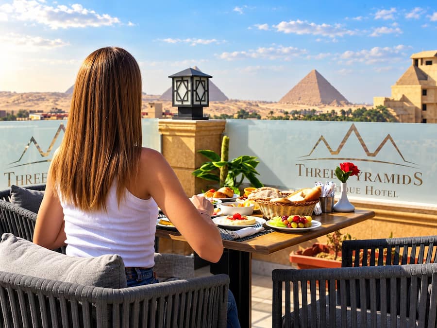 Une personne est assise à une table, devant laquelle on peut admirer les pyramides, et à proximité se trouvent une lampe et un panneau indiquant l'hôtel « Les Trois Pyramides ».