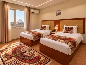 A hotel room with two beds, a large window with a view of buildings, an air conditioning unit, and a decorative rug. - Three Pyramids Boutique Hotel (Giza)