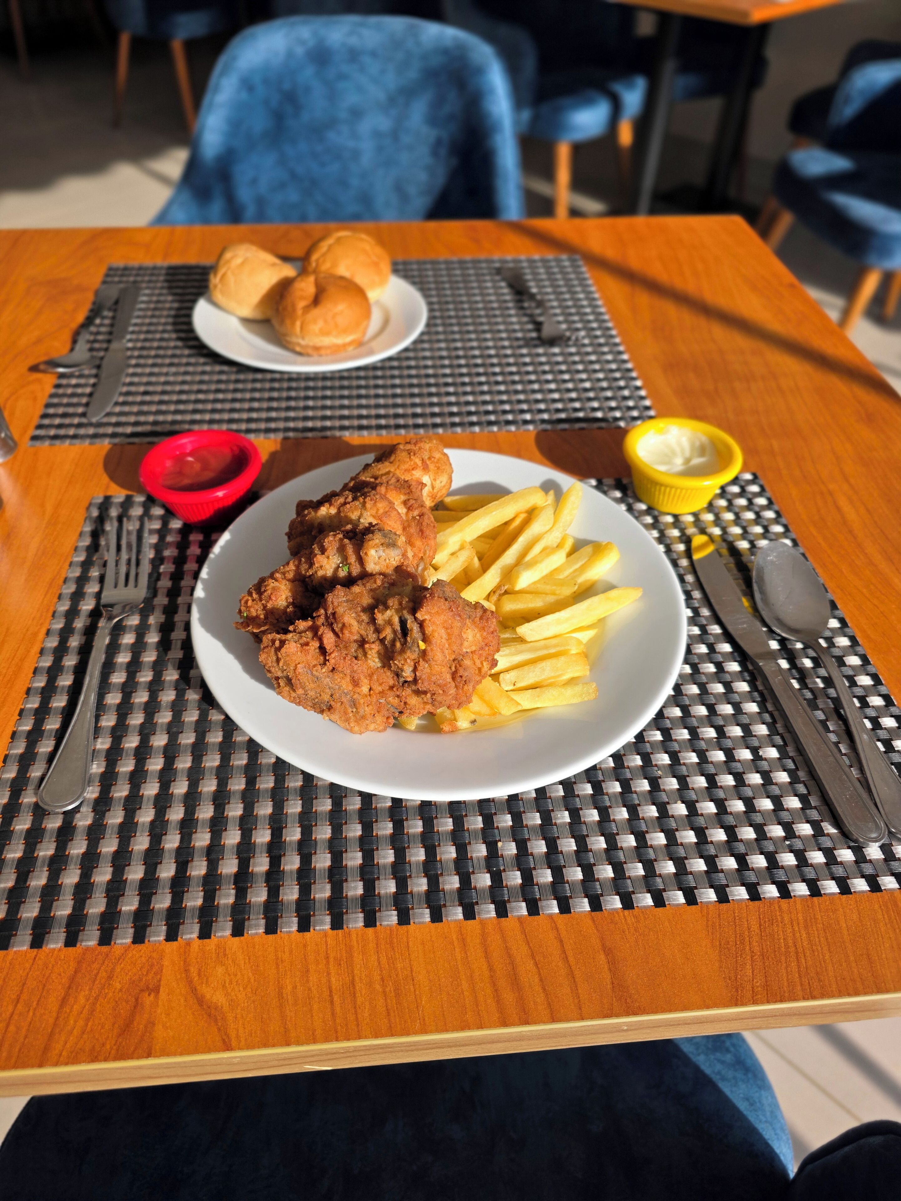 A dining table with a plate of fried chicken and fries, a small bowl of sauce, and a plate of rolls.