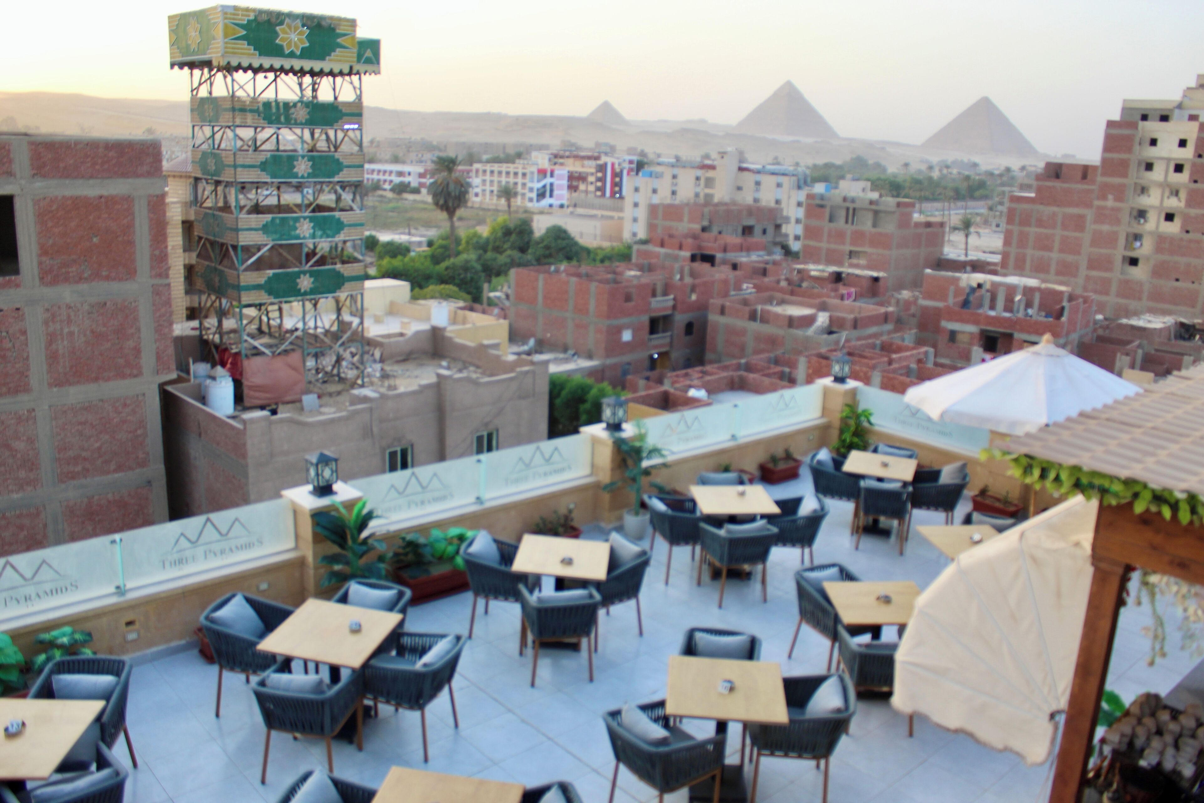 Una terraza exterior con varias mesas y sillas, vista a las pirámides en el fondo, y un edificio en construcción con andamios.