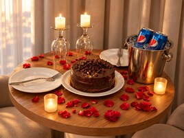 Un tavolo rotondo in legno con una torta al cioccolato, due candele accese, una lattina di Pepsi e petali di rosa sparsi.