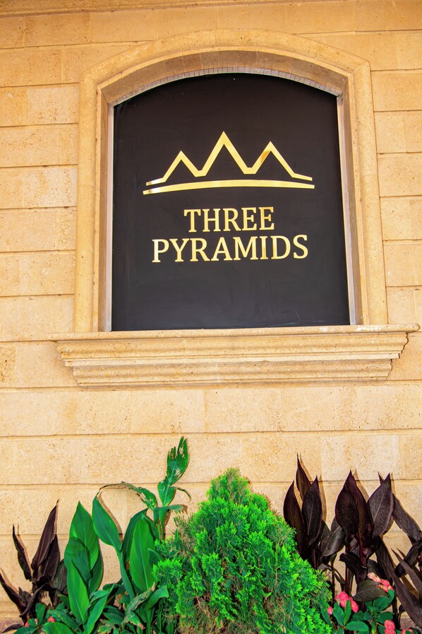 A sign with "THREE PYRAMIDS" in gold lettering on a black background, framed by a stone wall and surrounded by plants. - Three Pyramids Boutique Hotel (Giza)