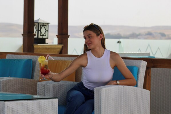A person seated on a wicker sofa with a view of a body of water and hills in the background. - Three Pyramids Boutique Hotel (Giza)