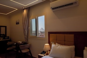 A hotel room with a bed, a desk with a chair, a lamp, an air conditioner, and a window with a view of buildings. - Three Pyramids Boutique Hotel (Giza)