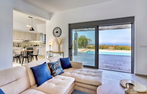 A modern living room with a large sofa, a dining area, and a view of a pool and landscape. - Sternes Panorama Executive Villa with Pool, Sitia (Sitia)