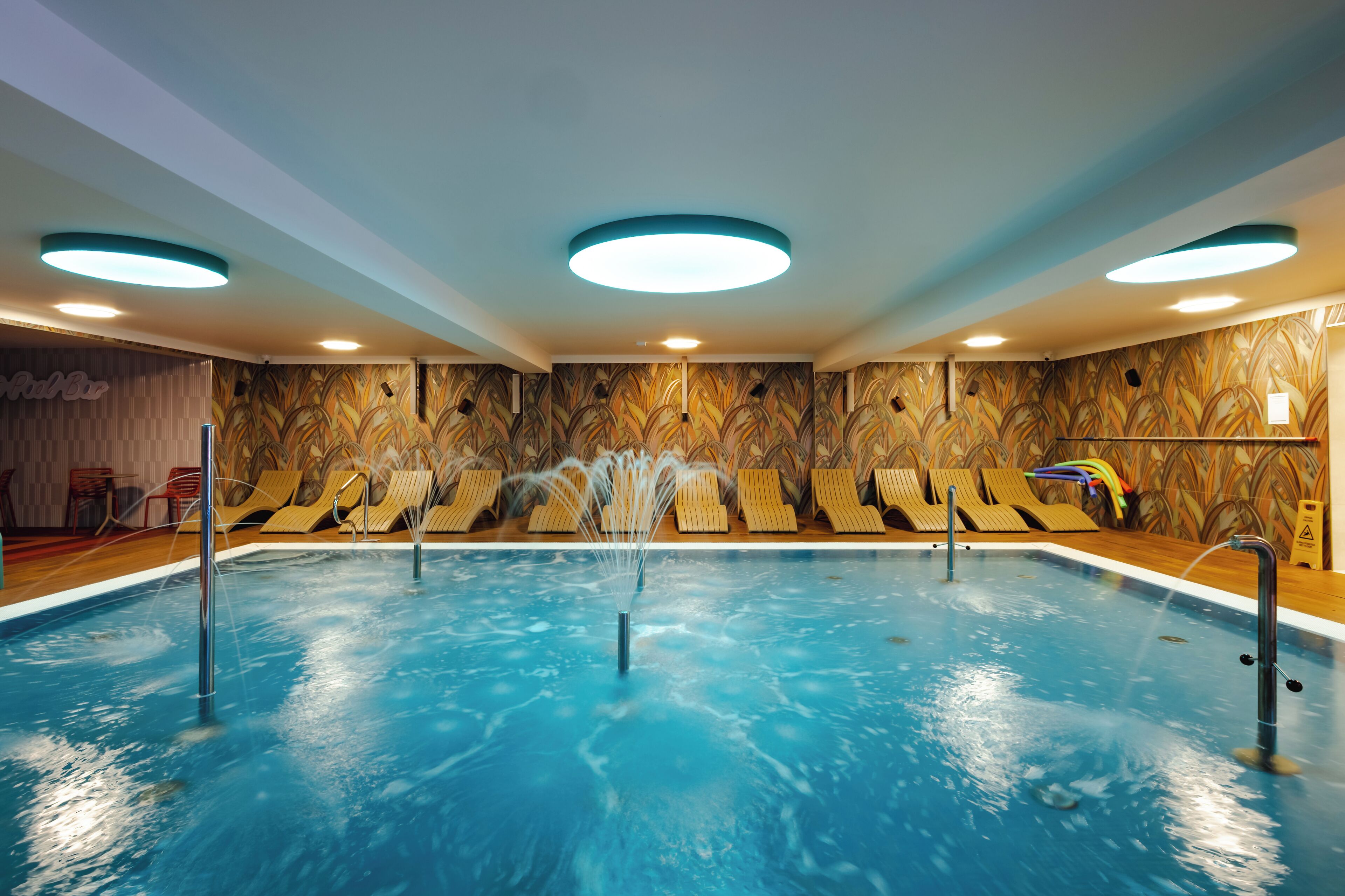 An indoor swimming pool with multiple lanes, lounge chairs, and a wall with a leaf pattern.