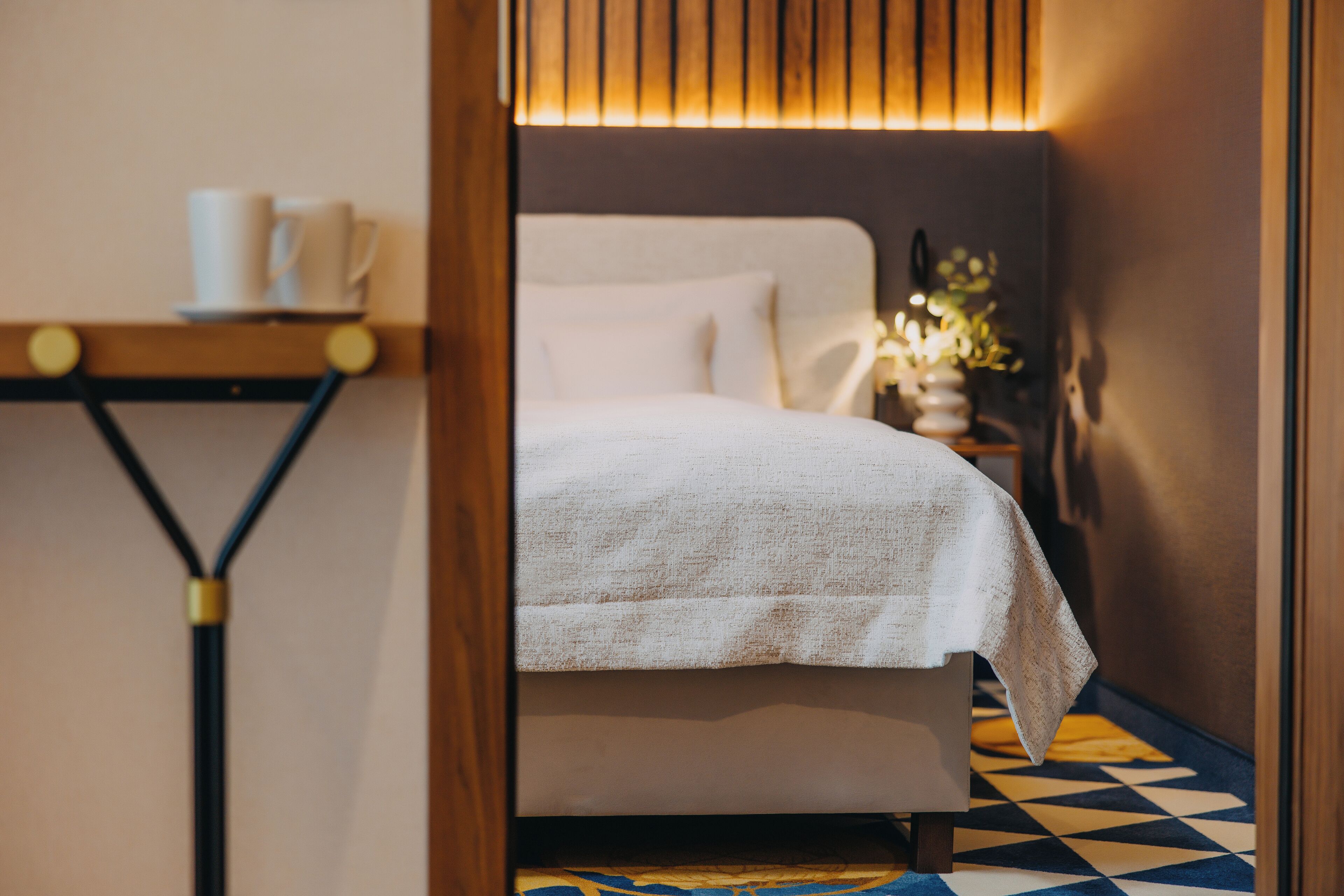 A neatly made bed with a white bedspread, a bedside table with a lamp, and a wall-mounted shelf with cups.