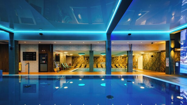 A modern indoor swimming pool with blue lighting, a reception area, and a seating area with chairs and a small pool.