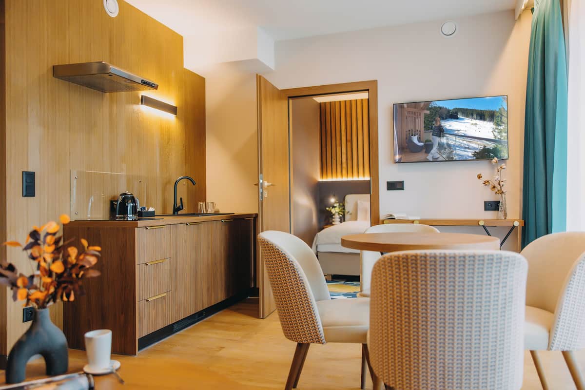 Una habitación de hotel moderna con una kitchenette de madera, una zona de comedor con dos sillas, un televisor montado en la pared y vistas a un paisaje nevado.