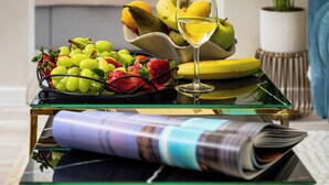 Ein Esstisch mit Obstschale, einem Weinglas und einer Zeitschrift. Im Hintergrund sind ein Bett und eine Pflanze zu sehen.
