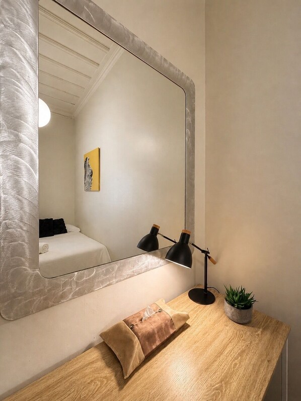 A room with a large mirror, a bed, a bedside table with a lamp, a small plant, and a pillow.