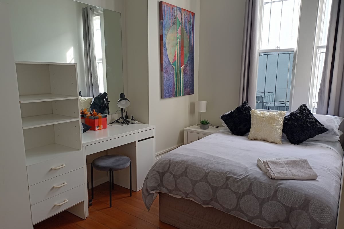 A bedroom with a bed, a desk, a chair, a large mirror, a painting, and a window.