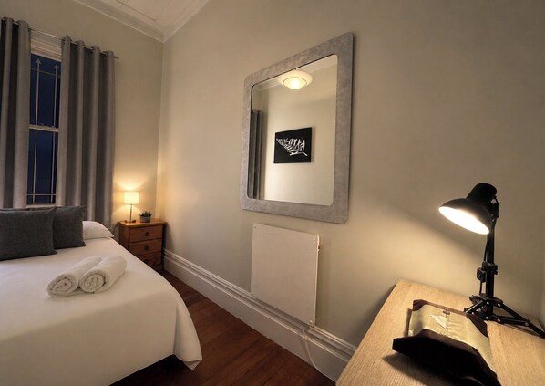 Une chambre d’hôtel comprenant un lit, une lampe de chevet, un miroir fixé au mur, une petite table avec une lampe et un radiateur.