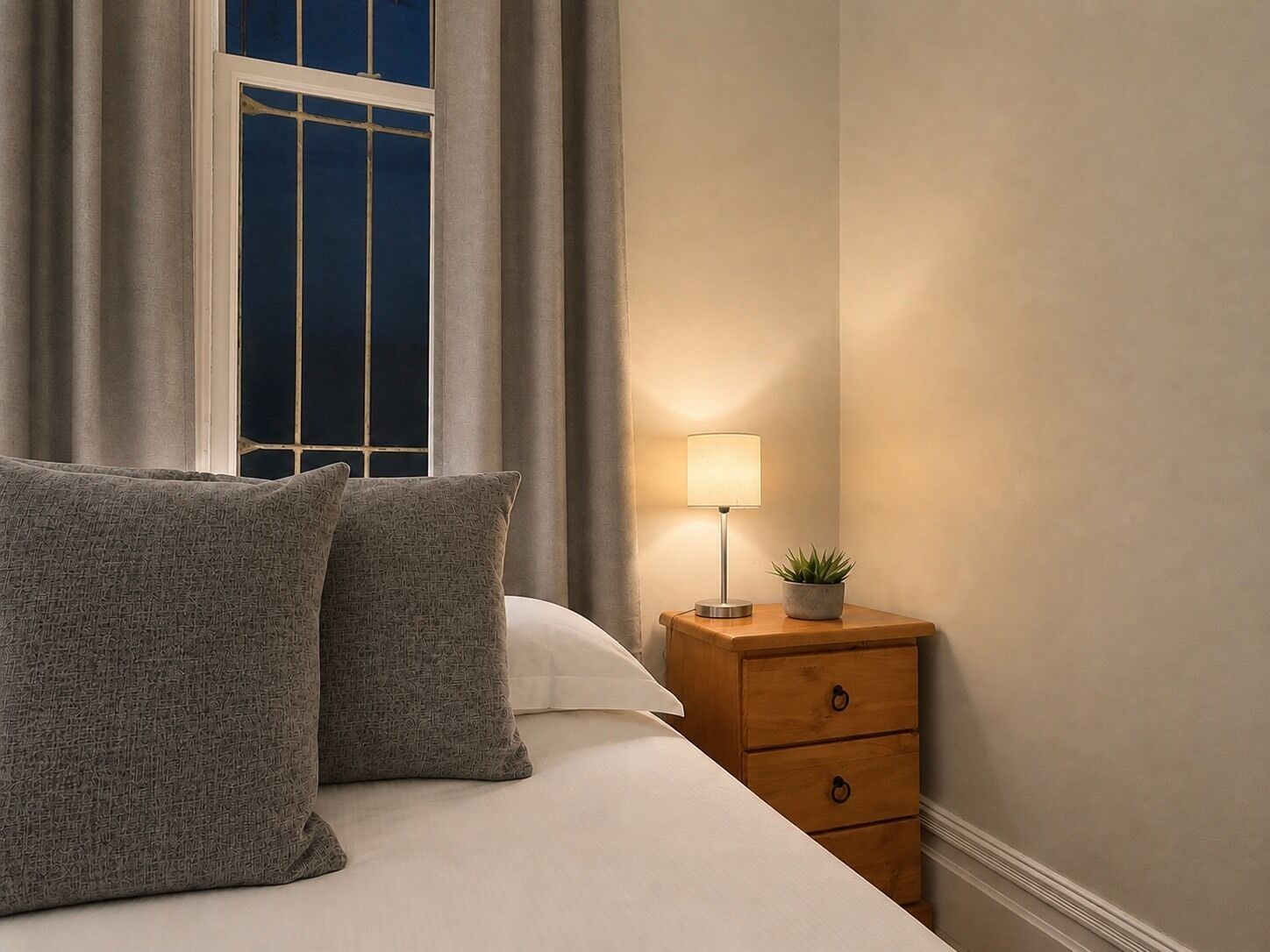 A neatly made bed with two pillows, a bedside table with a lamp and a small plant, and a window with curtains.