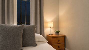 A neatly made bed with two pillows, a bedside table with a lamp and a small plant, and a window with curtains.