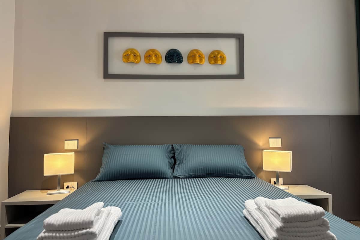 A hotel room with a bed, two bedside lamps, a framed artwork of masks, and neatly folded towels.