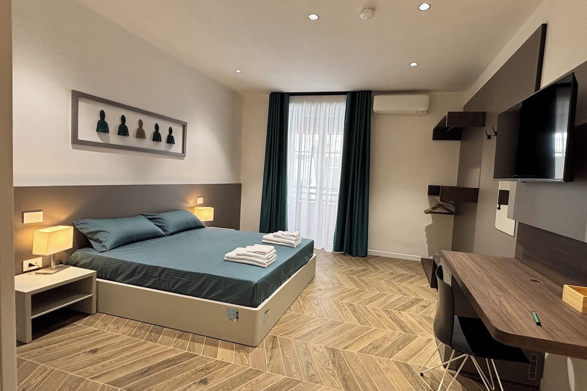 A modern hotel room with a bed, bedside lamps, a desk, a TV, and a herringbone-patterned floor.