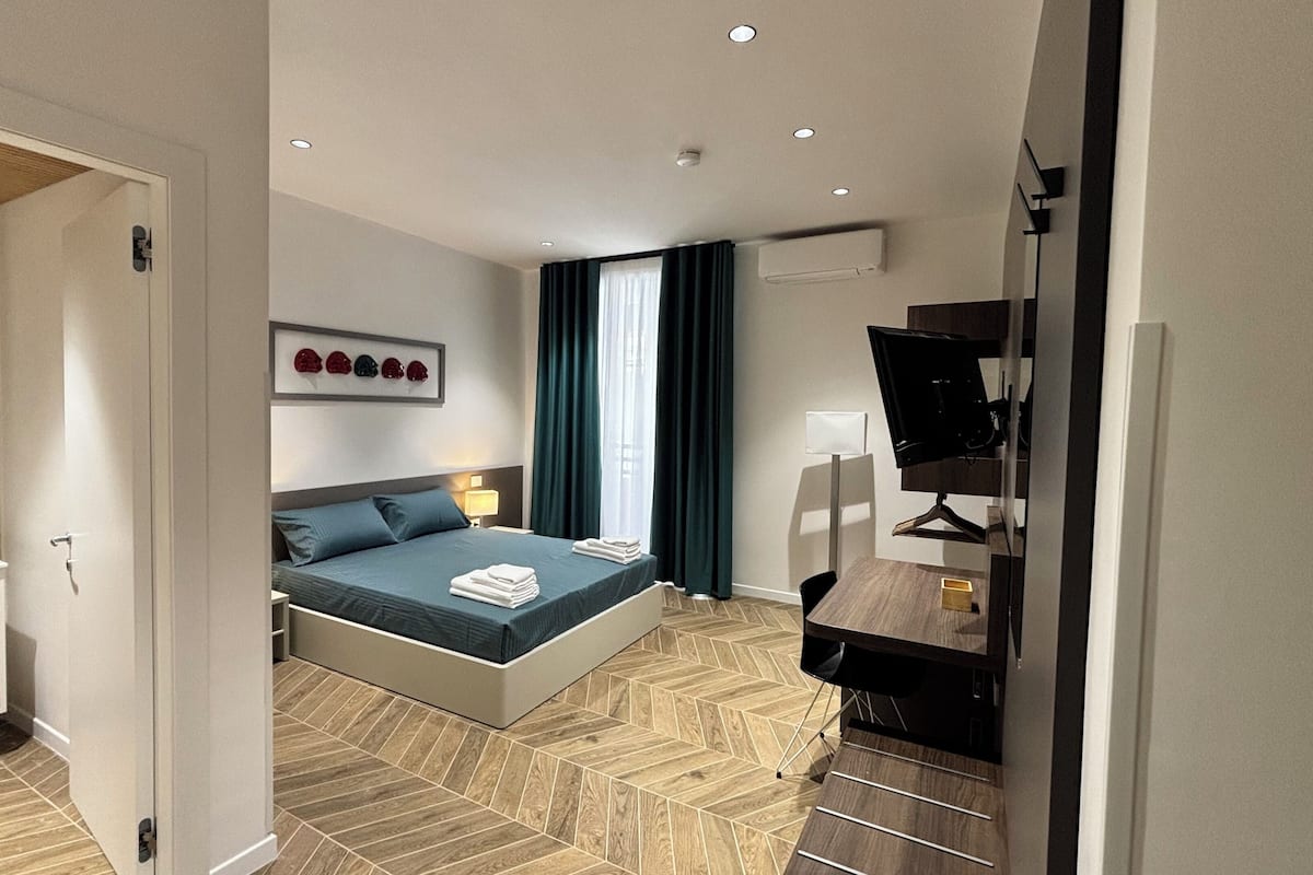 A modern hotel room with a bed, bedside table, lamp, and a desk with a computer monitor.
