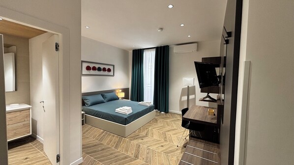 Ein modernes Hotelzimmer mit Bett, Nachttisch, Lampe und Schreibtisch mit Computerbildschirm.