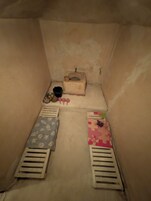 Ein kleiner, schummrig beleuchteter Raum mit einem Einzelbett, einem Tisch mit Eimer und Waschbecken sowie einer Rätscheldose auf dem Boden.
