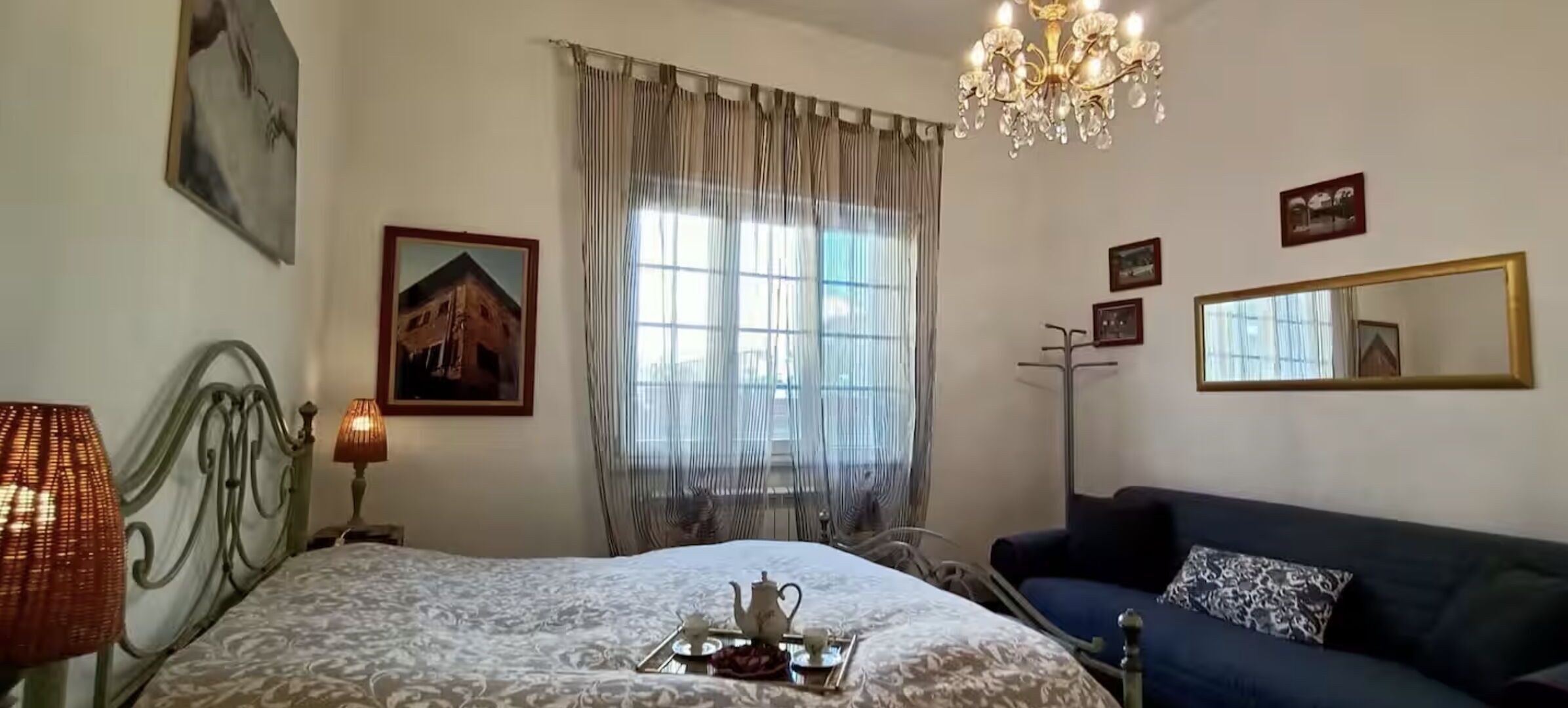 A bedroom with a bed, bedside lamps, a dresser, a blue sofa, a chandelier, a window with curtains, and wall-mounted pictures.