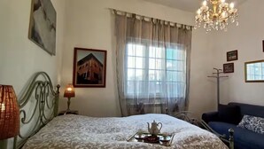 Une chambre à coucher avec un lit, des lampes de chevet, une commode, un canapé bleu, un lustre, une fenêtre avec des rideaux et des tableaux accrochés au mur.