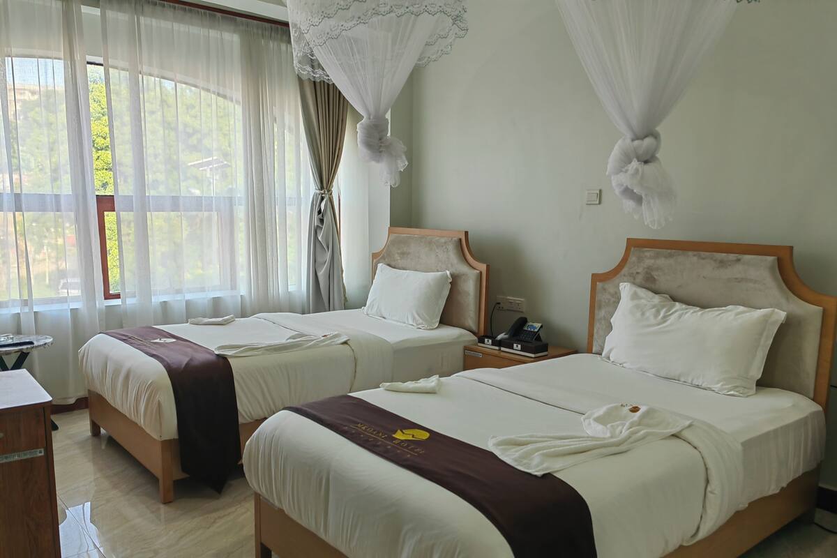 A hotel room with two beds, a bedside table with a phone, and a window with curtains.