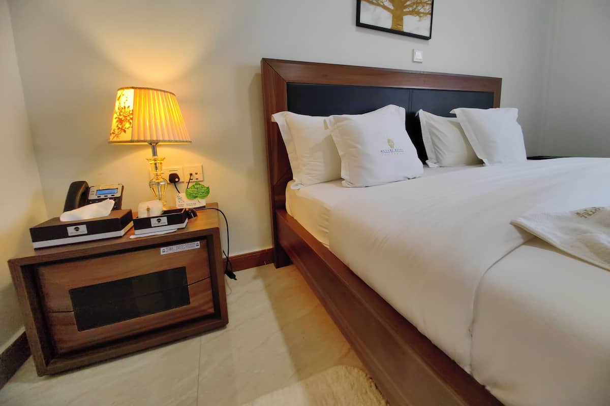 A neatly made bed with white linens and pillows, a bedside table with a lamp, a phone, and a small plant.