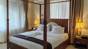 Ein Bett mit Baldachin, weiße Bettwäsche, ein Nachttisch mit Lampe sowie ein Beistelltisch mit Telefon und einer Schachtel.