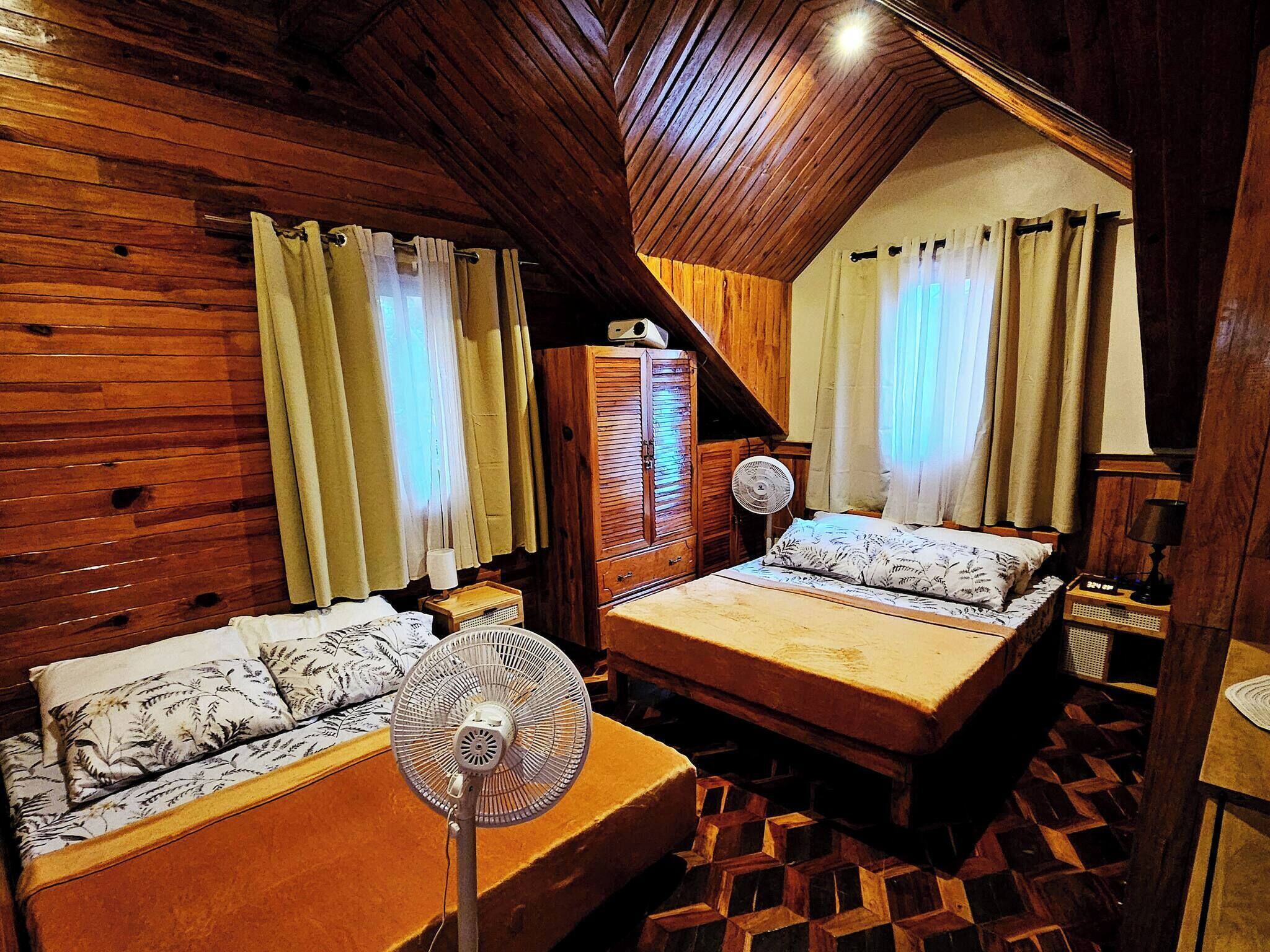 Una habitación acogedora con paredes de madera, techo inclinado, dos camas con ropa de cama floral y un ventilador de pie.