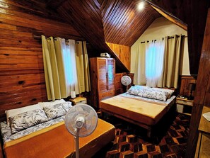 A cozy room with wooden walls, a pitched ceiling, two beds with floral bedding, and a standing fan.