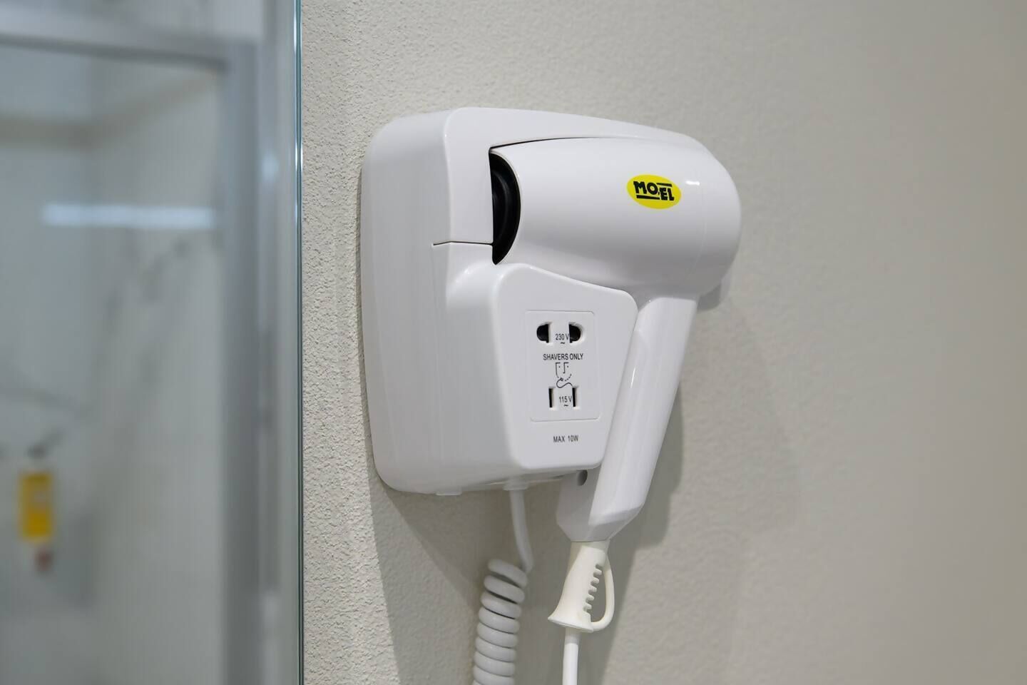 Ein weißer Haartrockner an der Wand montiert, mit einem gelben MOEL-Logo, einer 230-Volt-Buchse und einer 115-Volt-Buchse.