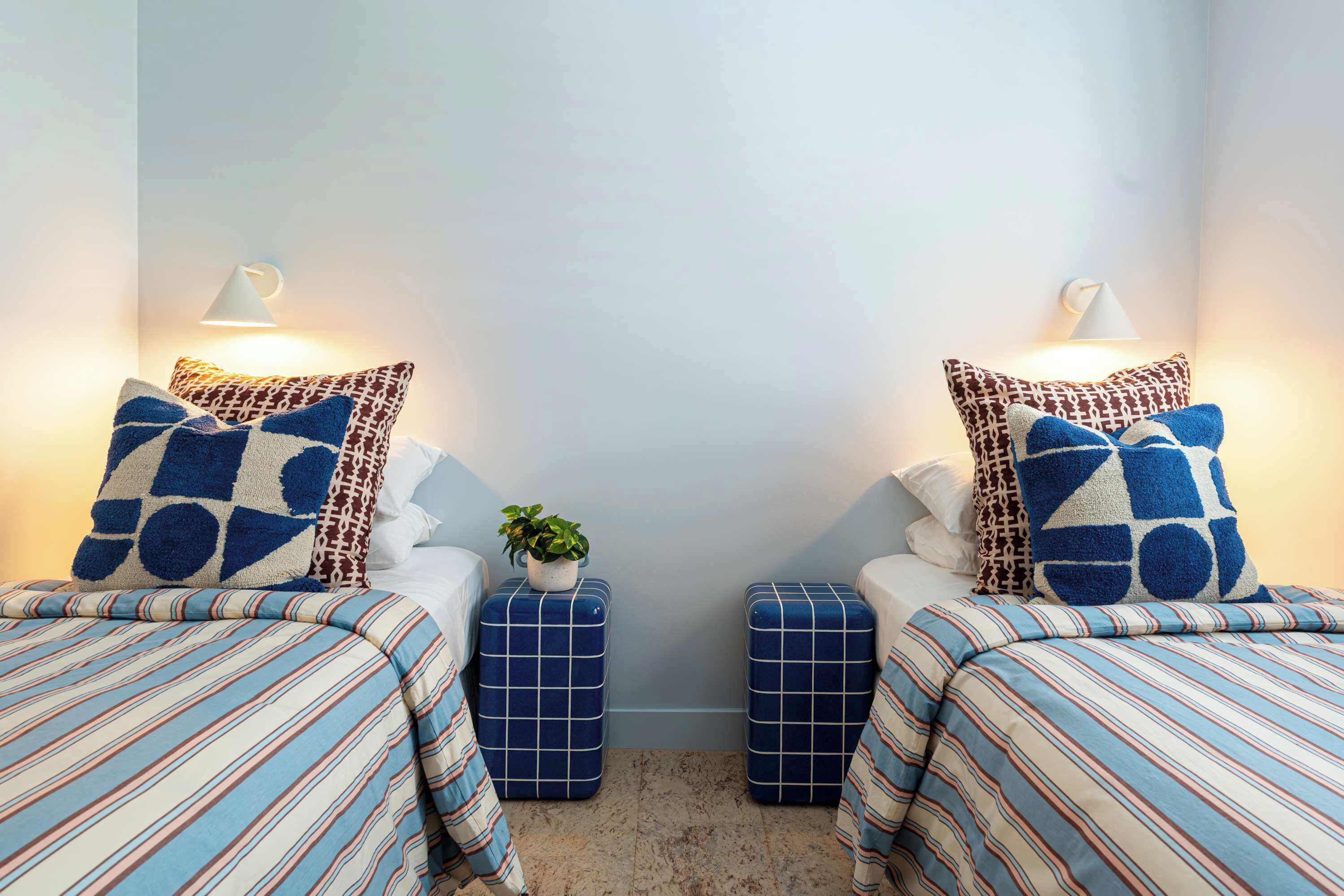 Una habitación con dos camas, cada una con ropa de cama y almohadas estampadas, una mesita con una planta y lámparas fijadas a la pared.