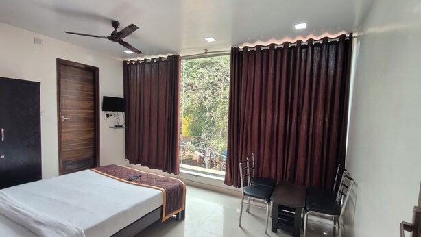 A bedroom with a bed, a desk, two chairs, a wardrobe, a ceiling fan, and a window with curtains. - Hotel Golden Arch  (Bangalore)