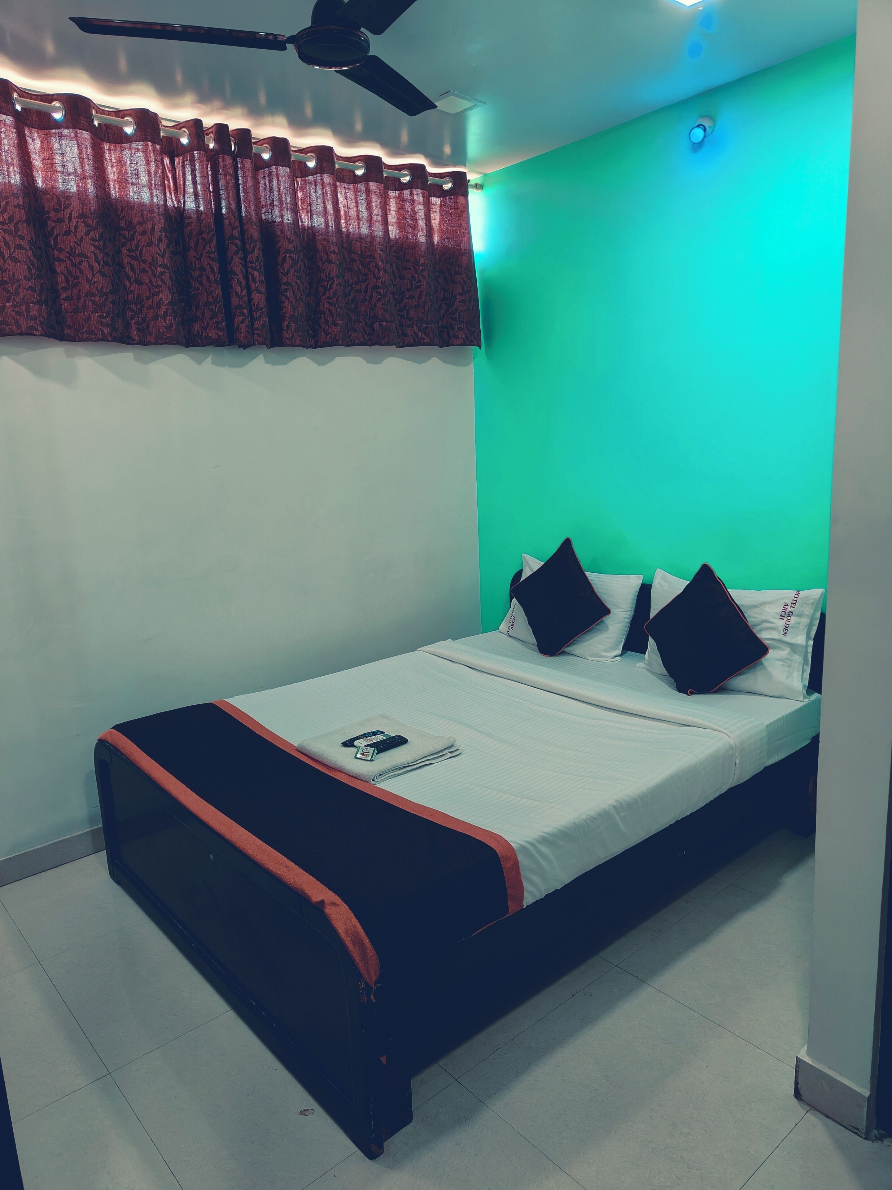A neatly made bed with a remote control and a folded towel, a ceiling fan, and a green wall.