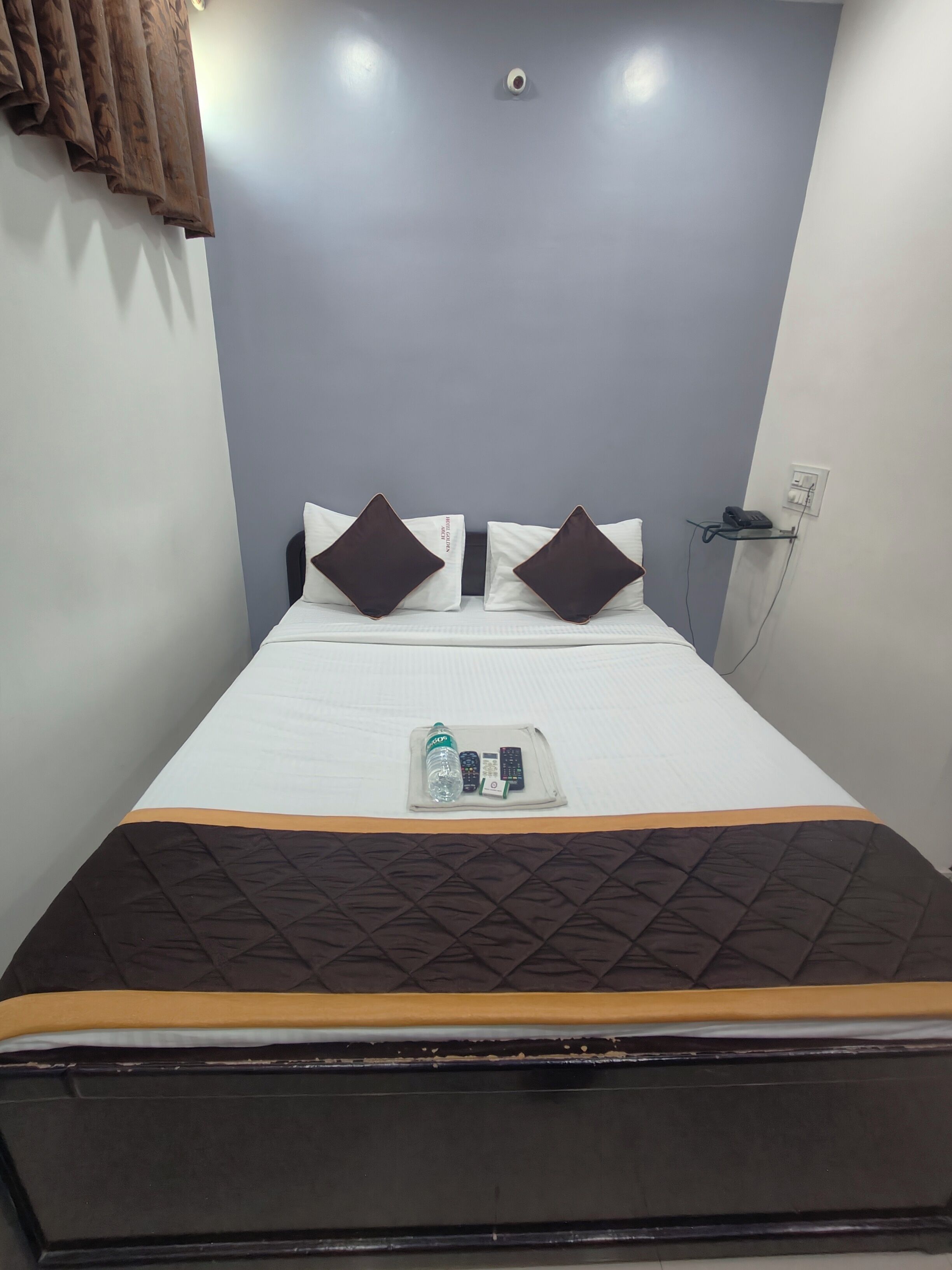 A neatly made bed with a quilted bedspread, two pillows, and a tray with a bottle and remote controls.